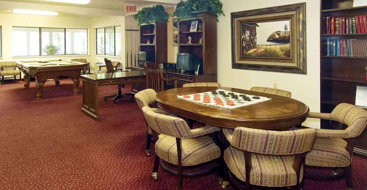 A cozy room featuring a table with a chessboard, surrounded by upholstered chairs, bookshelves, and decor.