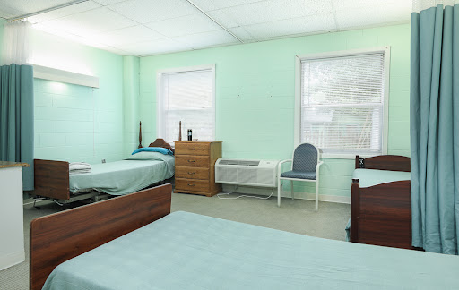 A bright and simple bedroom featuring two beds, a chair, and a dresser, with light green walls providing a calming atmosphere.