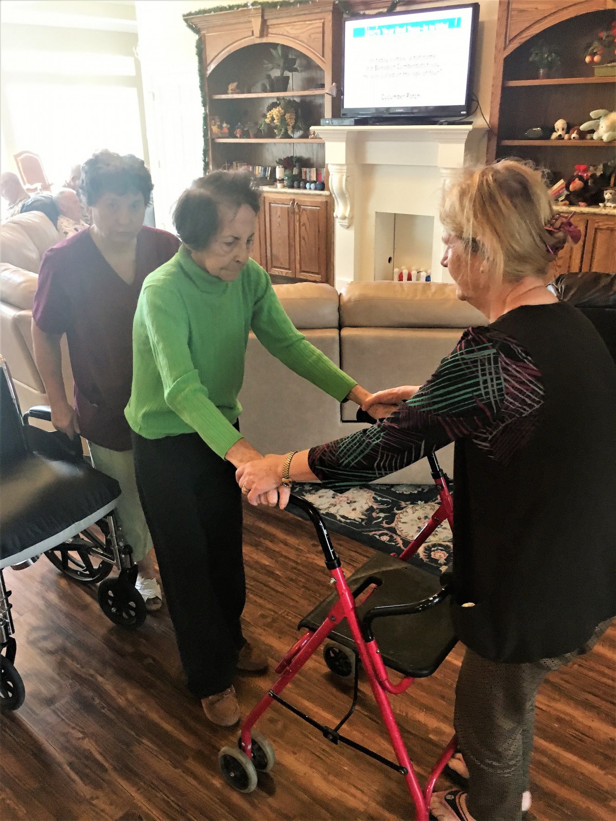 An elderly woman receives assistance from caregivers while using a walker in a nursing home setting.