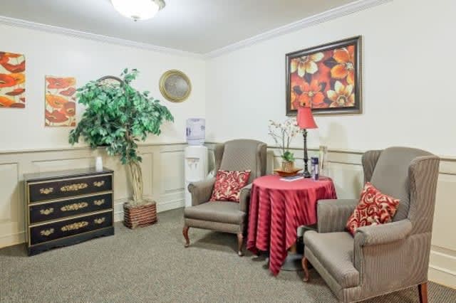 A cozy living room corner with two armchairs, a small table covered with a red cloth, and decorative elements including a lamp, wall art, and an indoor plant.