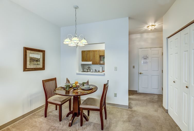 A cozy dining area with a round table and two chairs, featuring a light fixture above and a kitchen pass-through.