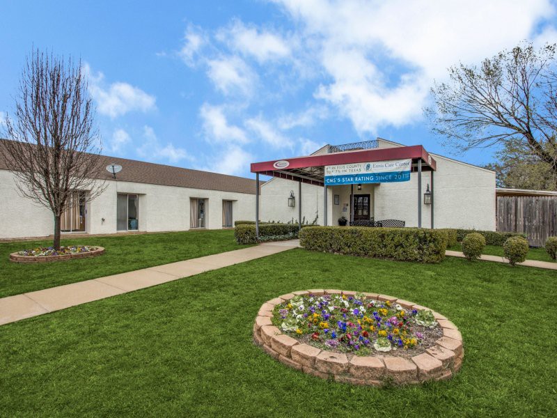 A building with a landscaped entrance featuring a flower bed and a green lawn on a sunny day.