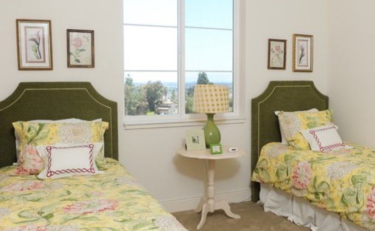 A cozy bedroom with twin beds featuring green headboards and floral bedding, separated by a side table with a lamp under a window.