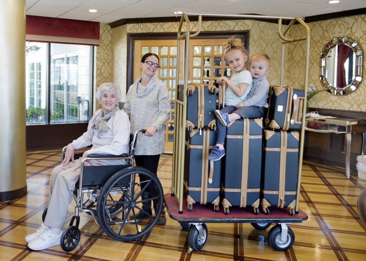 Children sit on a luggage cart while an elderly woman in a wheelchair and staff member smile in a hotel lobby.