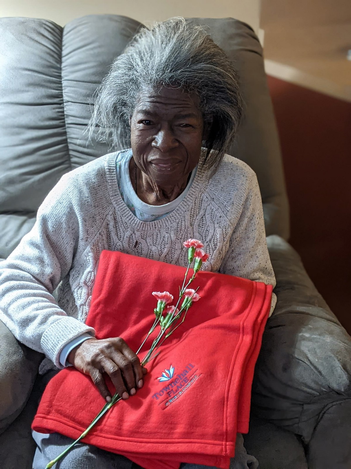 An elderly person holding flowers, seated with a red blanket in a cozy home setting.