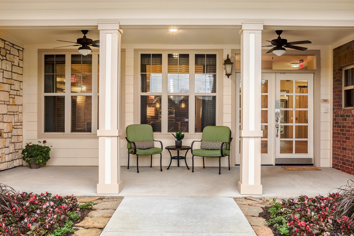 A welcoming porch with green chairs and a small table, framed by columns and illuminated by ceiling fans and outdoor lighting.