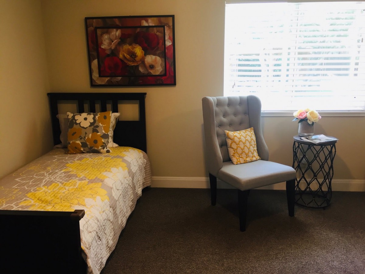 A cozy bedroom features a bed with floral bedding, a plush chair, and decorative elements like a painting and flowers.