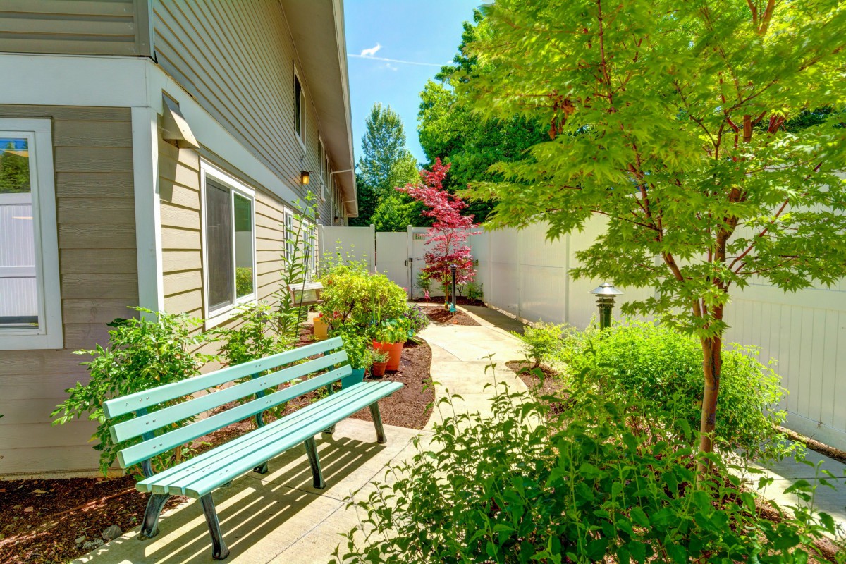 A serene garden with a bench and a pathway adjacent to a house, surrounded by lush greenery.