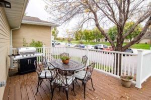 A cozy patio deck with a table, chairs, and a grill overlooking a neighborhood street.