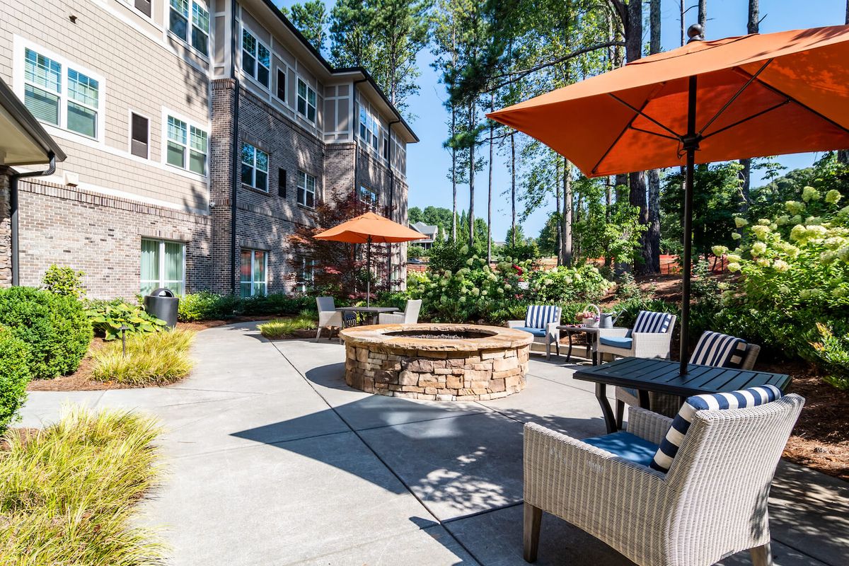 A cozy outdoor patio area with a fire pit and seating at an apartment complex, surrounded by lush landscaping and trees.