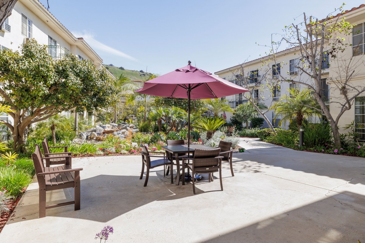 A sunny patio area featuring a table with chairs and a large umbrella, surrounded by a landscaped garden and residential buildings.
