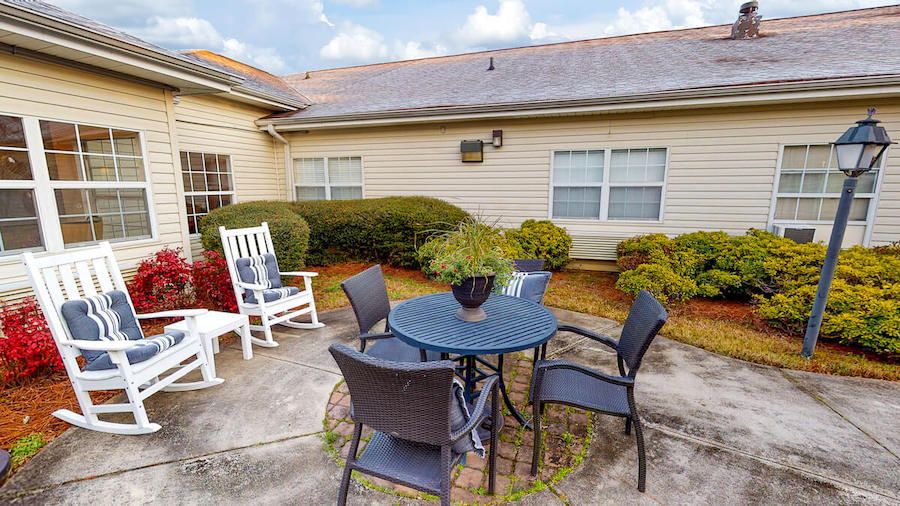 Outdoor patio area with a table, chairs, and a rocking chair surrounded by shrubs.