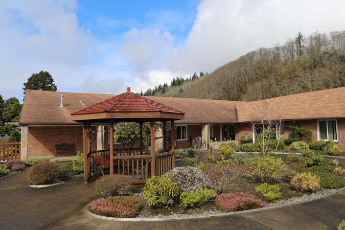 A peaceful courtyard featuring a wooden gazebo surrounded by landscaped gardens and a building in the background.