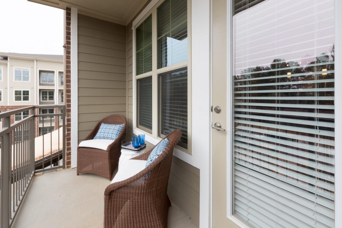 A cozy balcony with wicker chairs and cushions, next to a door and windows with blinds.