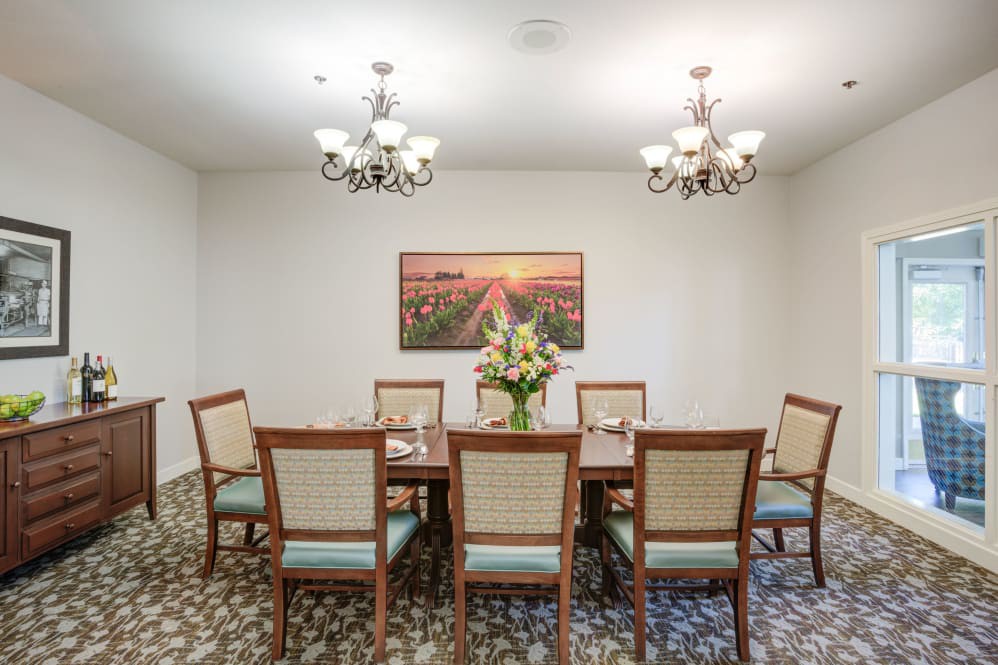 A well-lit dining room with a long table, surrounded by chairs and decorated with flowers and artwork.
