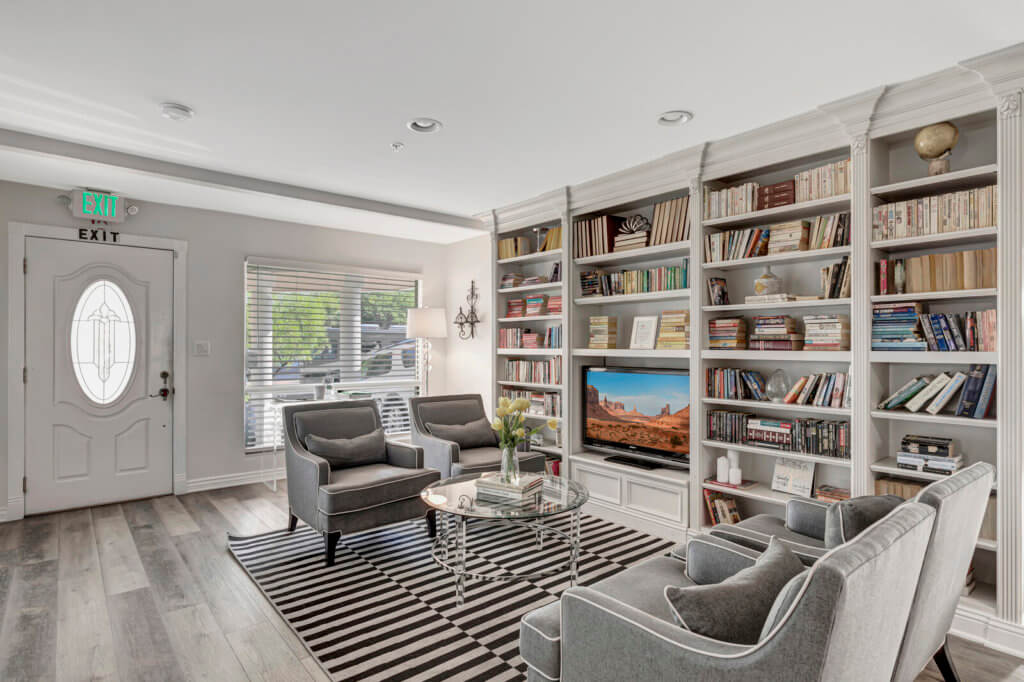 A modern living room featuring built-in bookshelves, comfortable armchairs, and a striped rug, with ample natural light streaming in through large windows.