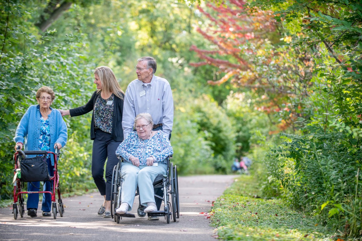 A family enjoying a walk in the park, with one member in a wheelchair, surrounded by lush greenery.