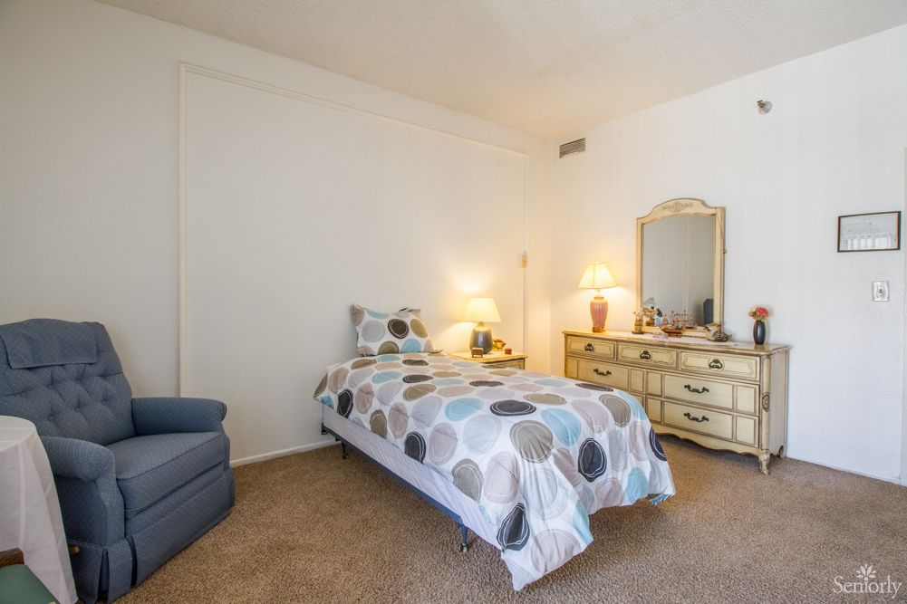 A cozy bedroom with a single bed, patterned bedding, dresser with mirror, and warm lighting.