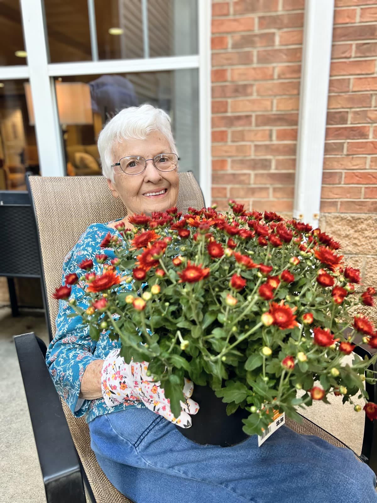 An elderly woman smiling while holding a large pot of vibrant flowers outdoors.