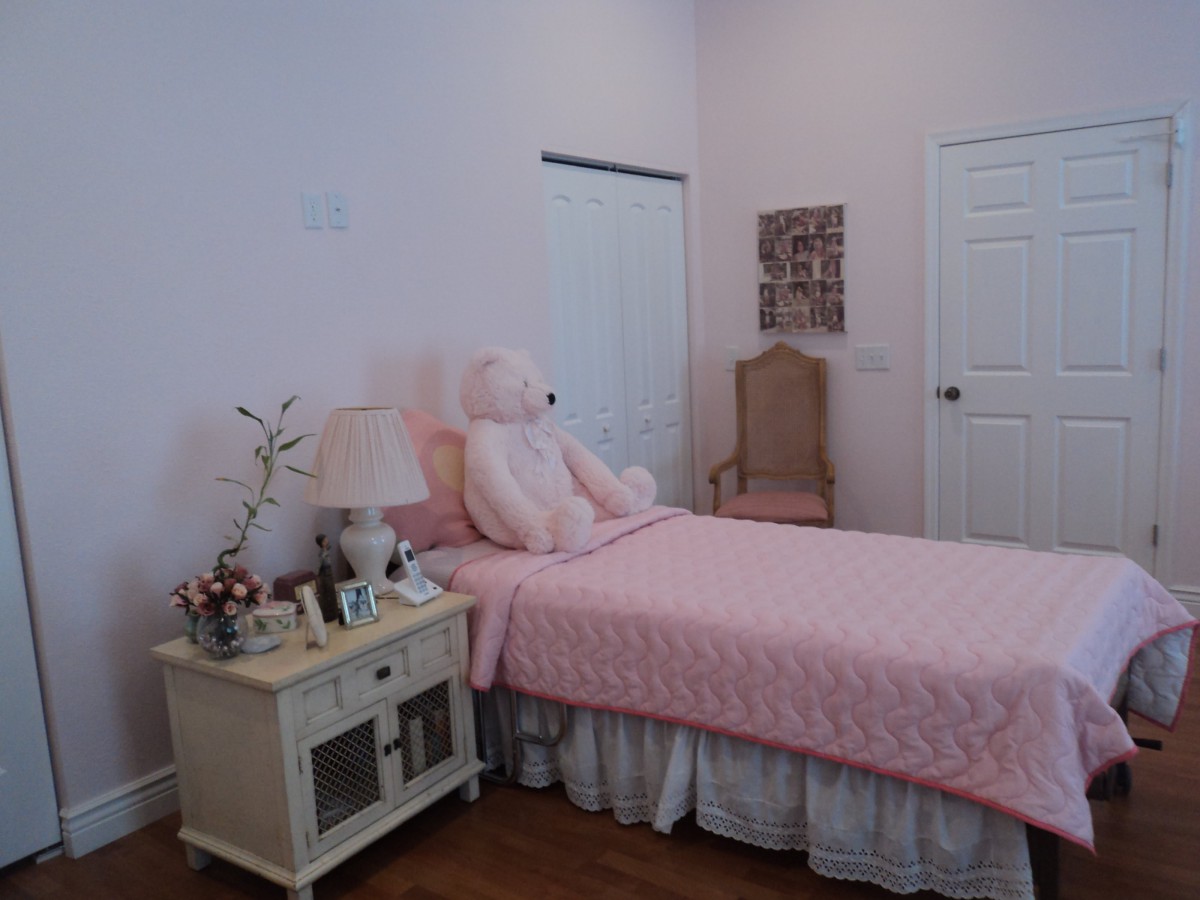 A cozy bedroom with pink decor, featuring a single bed adorned with a large teddy bear and a nightstand.