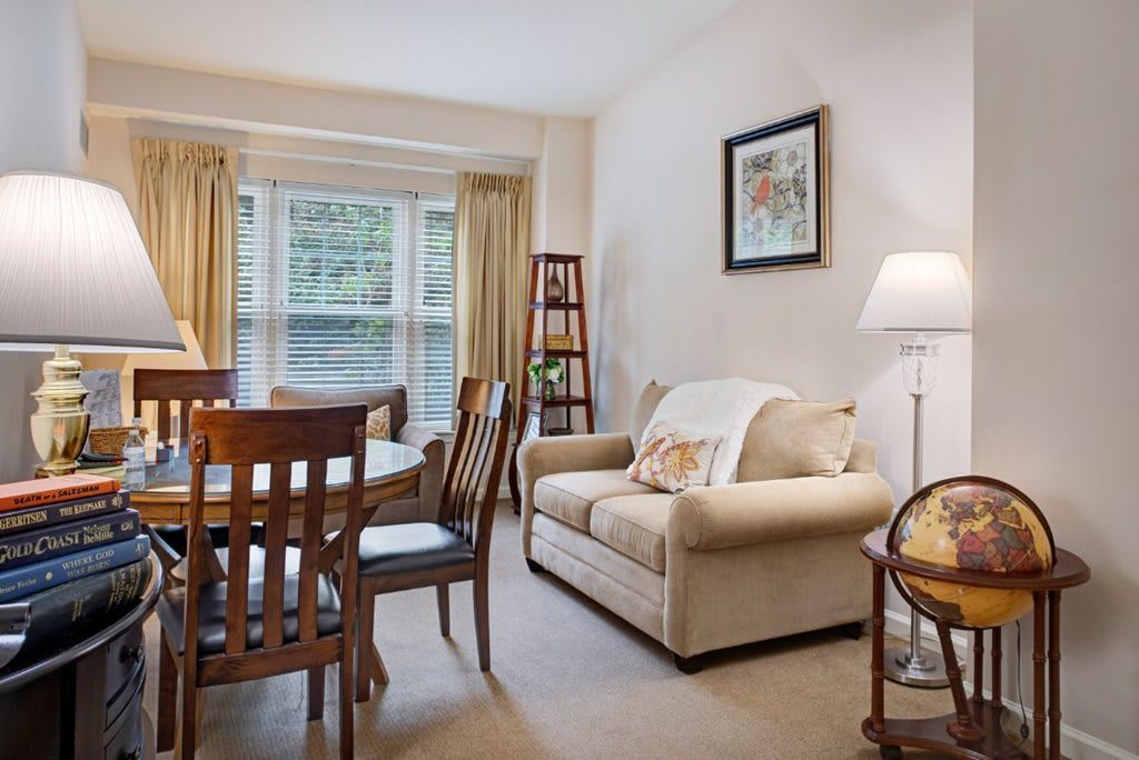 A cozy living room with a dining area, featuring a beige sofa, wooden dining table, and natural light from the window.