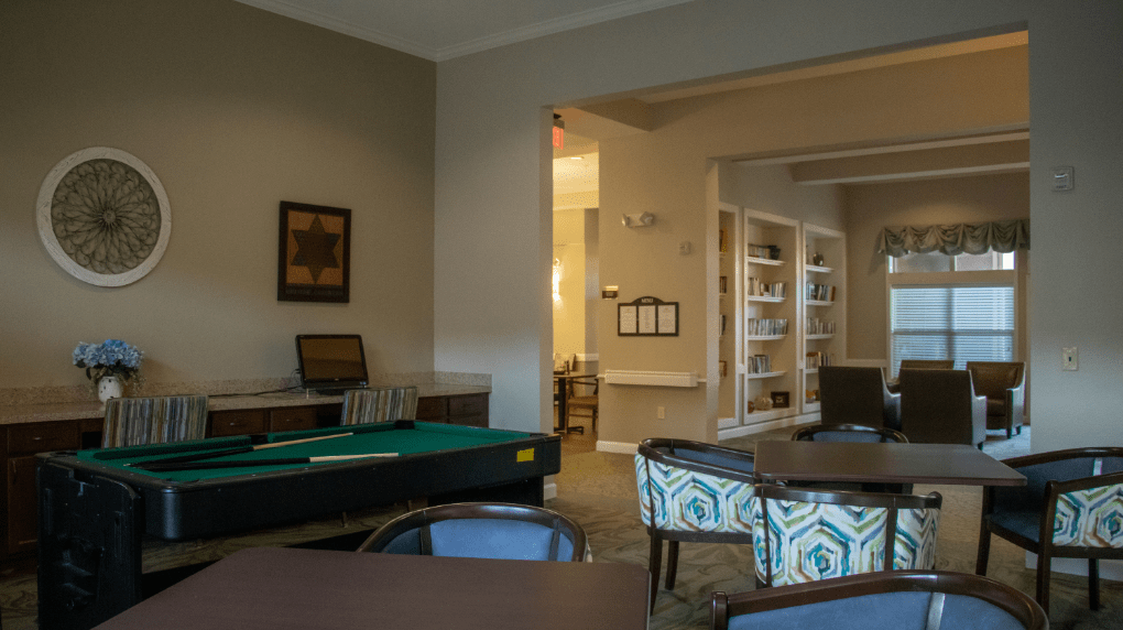 A cozy interior room with a pool table, tables and chairs, and bookshelves in the background.
