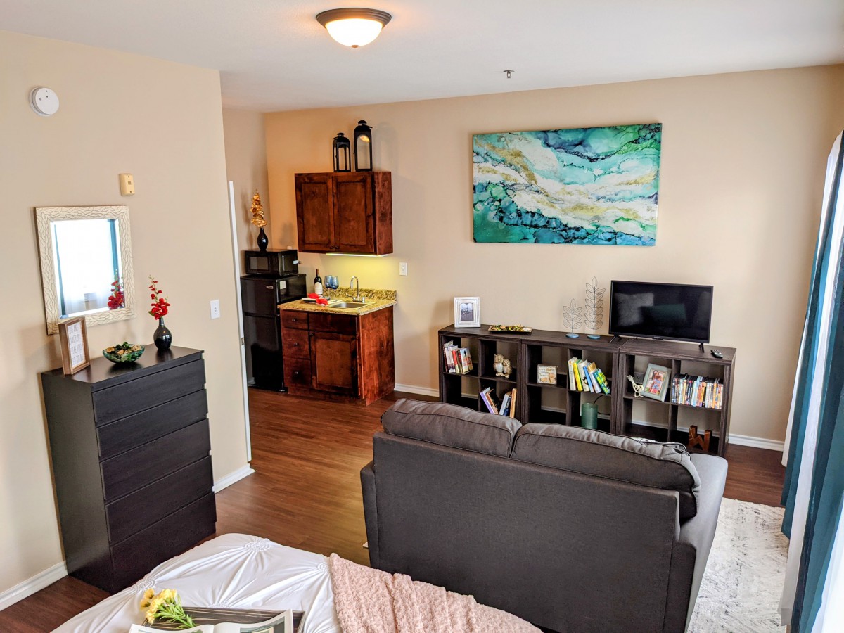 A cozy apartment living area with a view of the kitchen, featuring a sofa, artwork on the wall, and a television.
