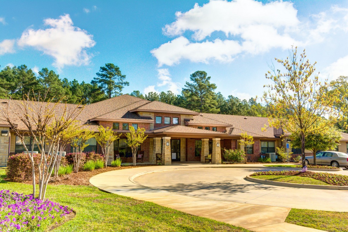 A beautiful residential building with a wide driveway, surrounded by trees and a sunny, cloudy sky.