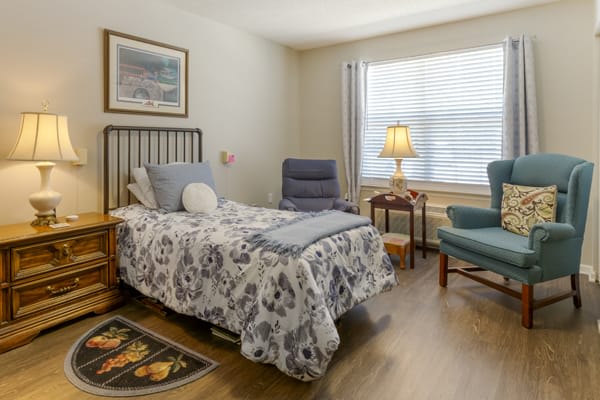A cozy bedroom featuring a bed with patterned bedding, a comfortable chair, and a lamp by the window.