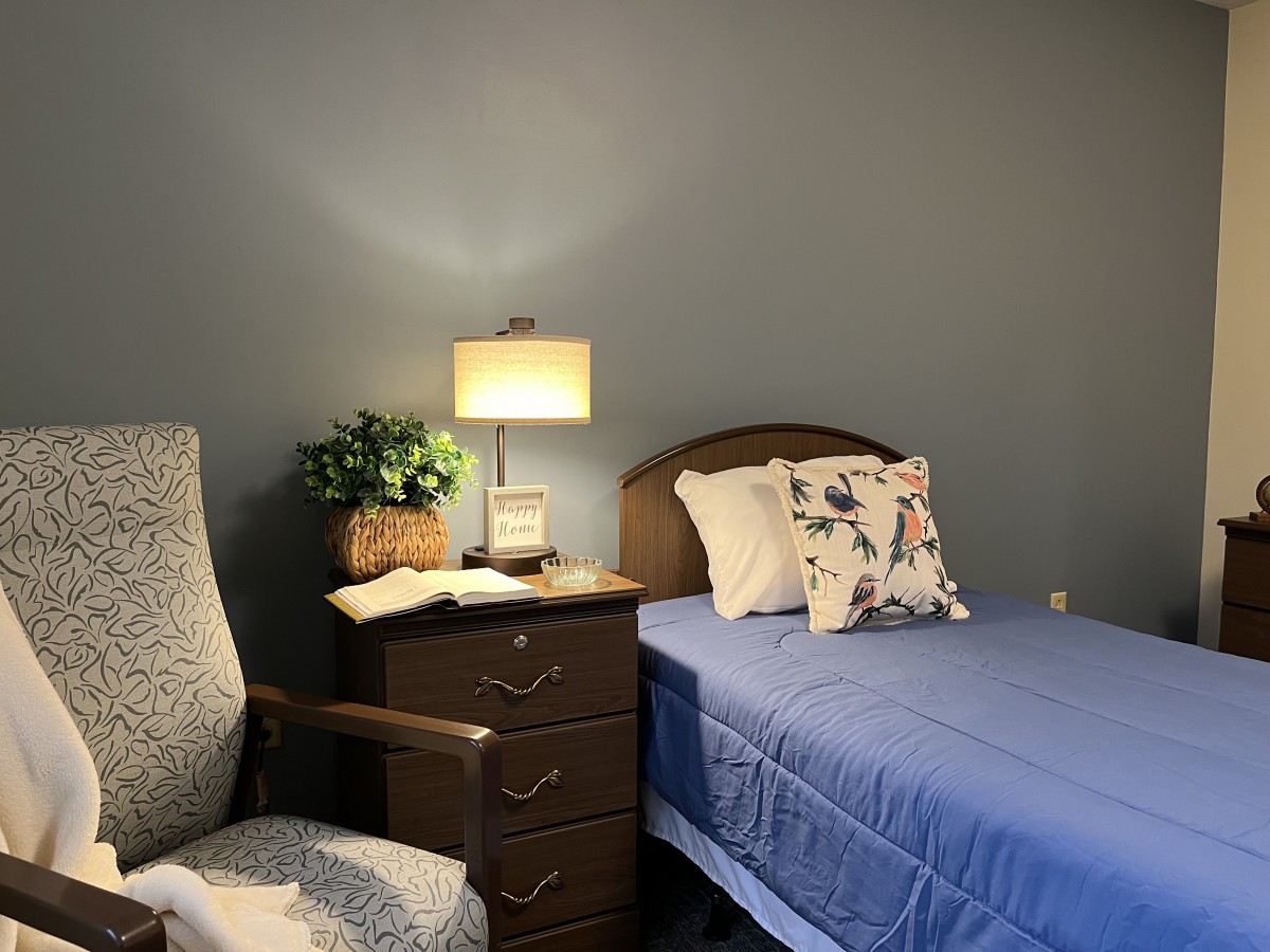 A cozy bedroom with a bed, nightstand with a lamp, and an upholstered chair.