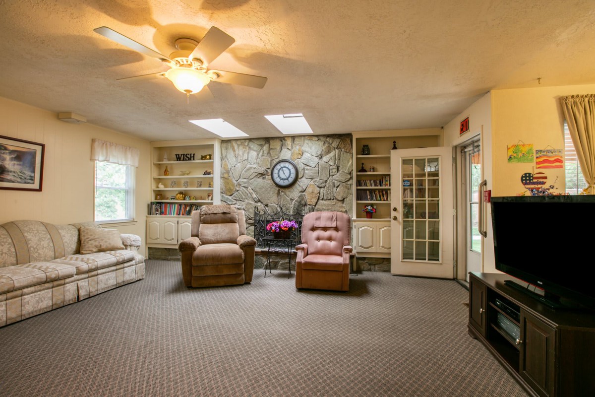 A cozy living room featuring comfortable armchairs, a soft carpet, ceiling fan, skylights, and built-in bookshelves surrounding a central stone fireplace.