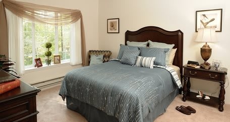 A cozy bedroom with a neatly made bed featuring blue bedding and several pillows, complemented by a large window allowing natural light to fill the space.
