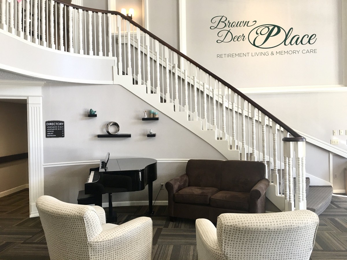 A cozy living room with a grand staircase, featuring comfortable seating and a piano.