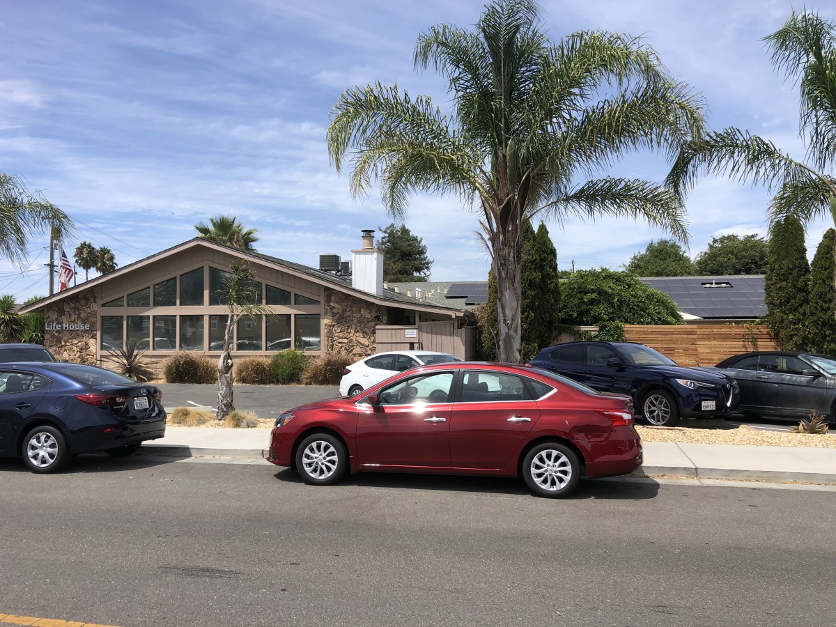 A suburban street with a palm tree, parked cars, and a house on a sunny day.