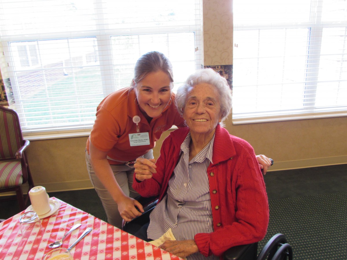An elderly woman in a wheelchair is smiling while posing with a caretaker at a dining table.