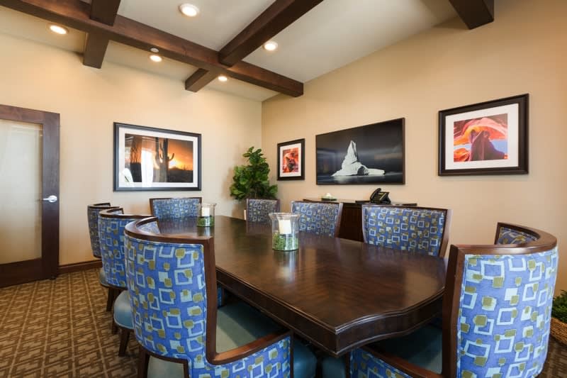 A modern conference room featuring a large wooden meeting table surrounded by upholstered chairs, with framed artwork on the walls and ceiling beams adding to the decor.