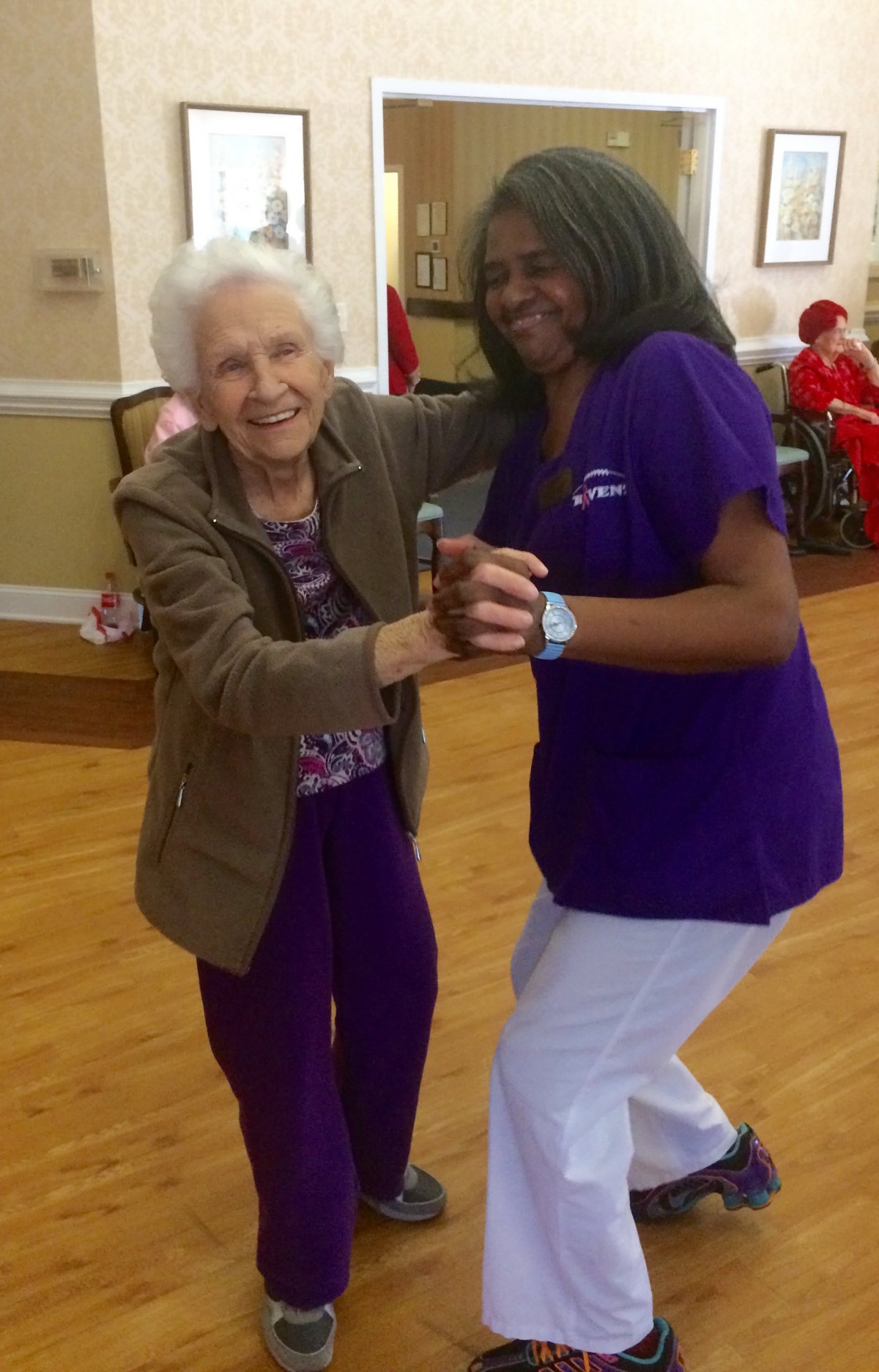 An elderly woman joyfully dances with a caregiver in a nursing home setting.