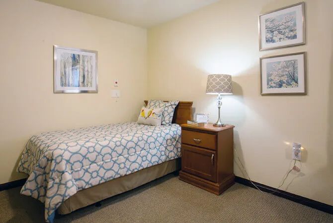 A cozy bedroom featuring a single bed with a patterned duvet, a wooden nightstand with a lamp, and framed artwork on the walls.