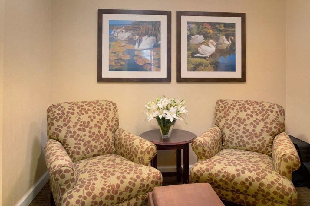 A cozy lobby area with two patterned chairs, a small table holding a vase of white flowers, and two framed paintings on the wall.