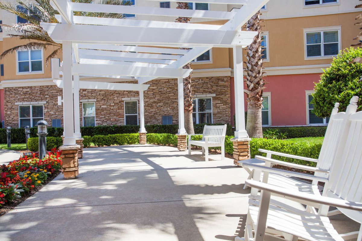 A peaceful outdoor seating area with a pergola providing shade, featuring wooden benches surrounded by landscaped greenery.