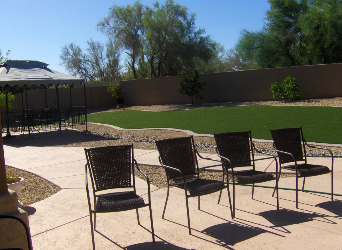 A sunny backyard with patio chairs arranged on a paved area, overlooking a grassy lawn enclosed by a fence.