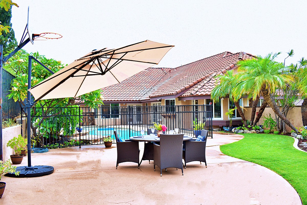 A spacious patio area with a dining set under a large umbrella, adjacent to a pool, with a house and lush greenery in the background.