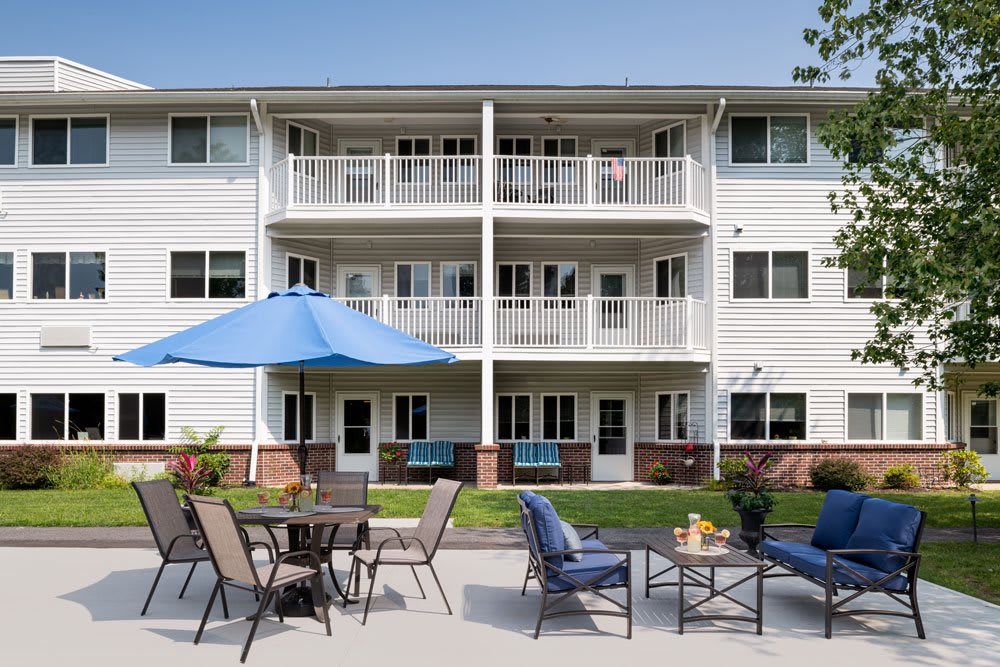 A three-story apartment building with balconies and an outdoor patio area featuring tables, chairs, and a large umbrella.