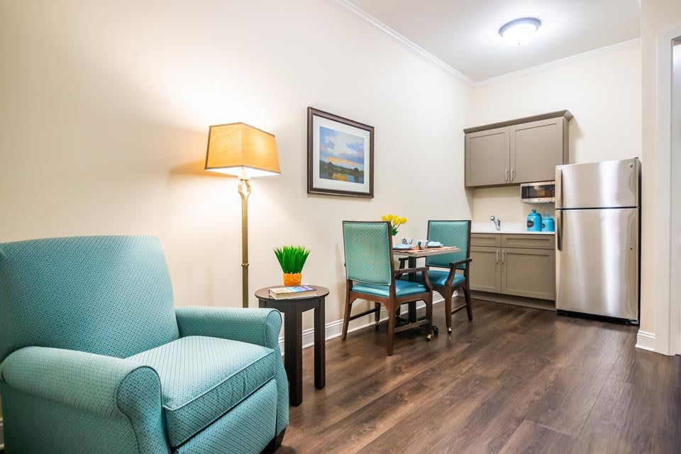 A cozy living room area with a light blue armchair, wooden flooring, and a kitchenette in the background.