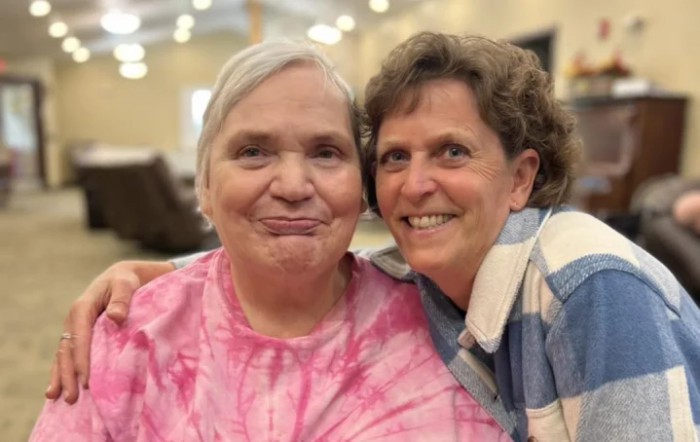 A middle-aged woman affectionately posing with an elderly woman indoors.