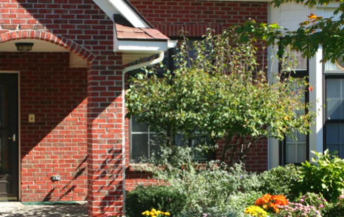 A charming brick house with a lush garden in front, featuring colorful flowers and a small tree.