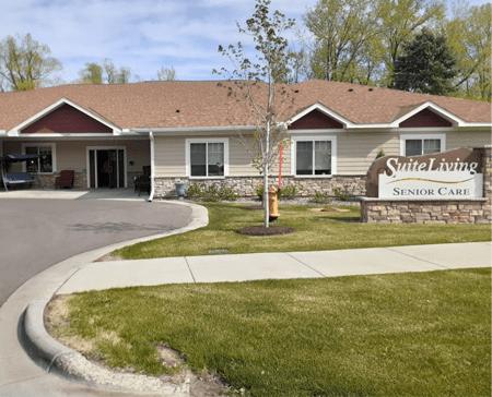 A single-story building with a driveway and a landscaped lawn featuring trees and a sign at the entrance.