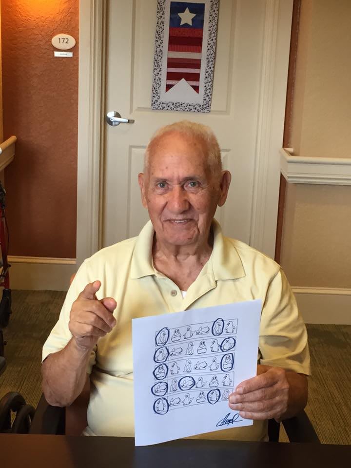 A senior man seated indoors, holding up a sheet of paper.