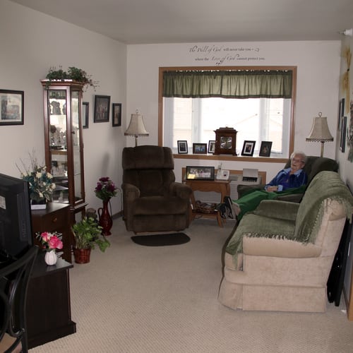 A cozy living room with a sofa, armchairs, and a television. The room is decorated with framed photos and plants.