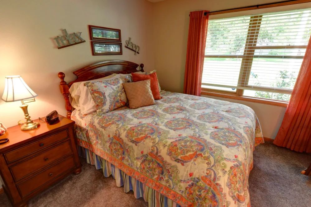 A cozy bedroom with a neatly made bed and patterned bedding, complemented by warm-colored curtains and natural light streaming through the window.
